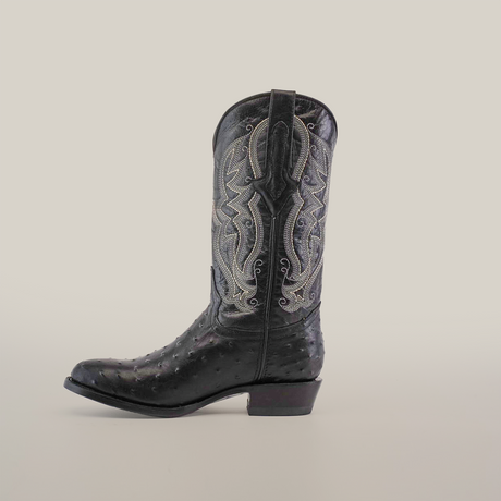 A side view of a single black Karoo Ostrich Print cowboy boot with a round toe, featuring decorative silver stitching and a textured pattern, crafted from cowhide leather, set against a plain white background.