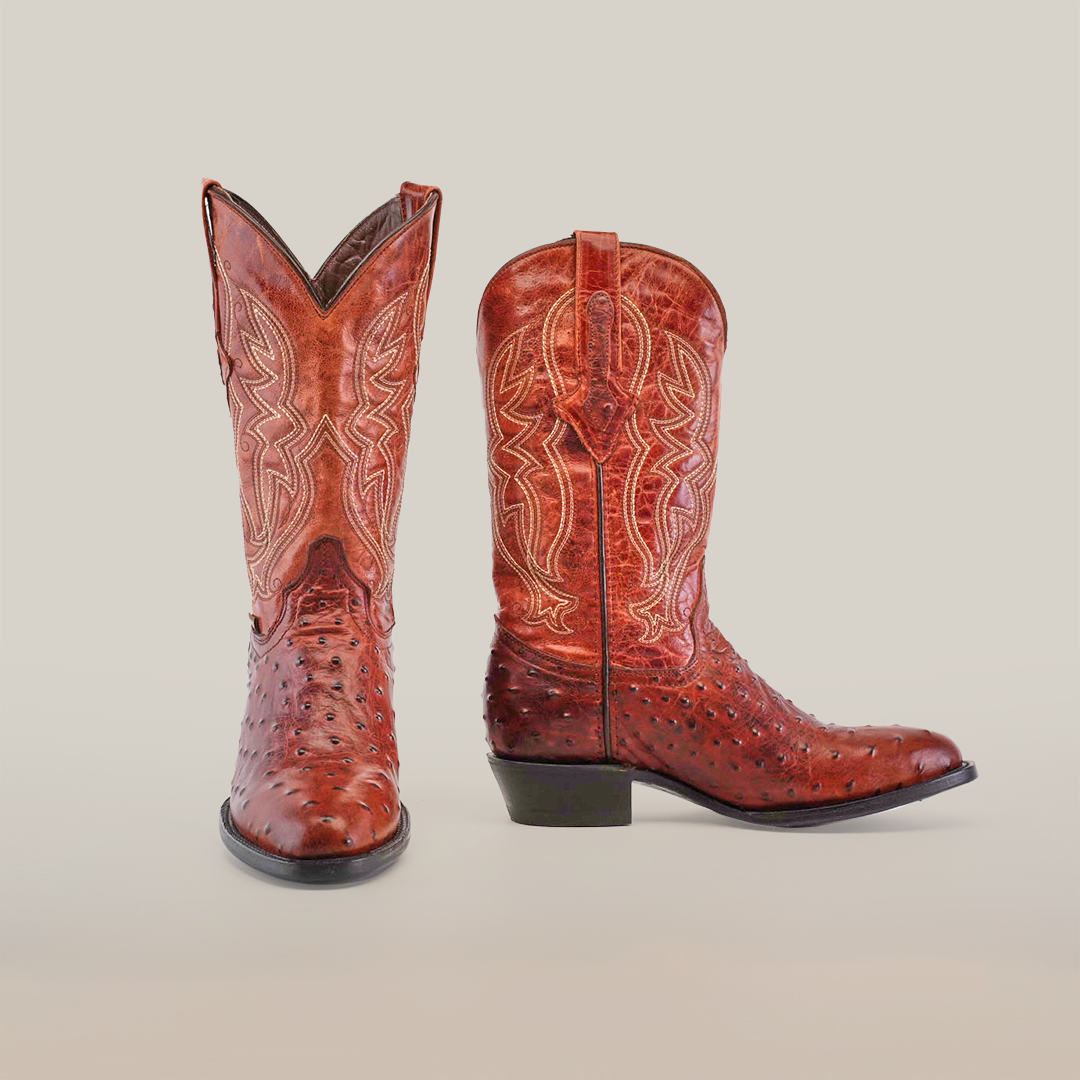 A pair of Karoo Ostrich Print Cogñac - Round Toe boots, crafted from premium cowhide with intricate stitching, stands against a plain white background. The left boot faces forward to highlight the detailed pattern, while the right boot angles to the side to emphasize its classic shape.