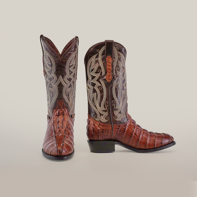 The Crocodile Tail Print Chocolate Round Toe cowboy boots are crafted from premium cowhide, showcasing intricate gray and tan stitching on the upper shafts. One boot is shown facing forward to highlight the toe design, while the other shows off its profile with heel, side embroidery, and leather sole.