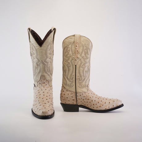 The Karoo Ostrich Print Bone boots, in a light color, feature intricate stitching and an ostrich print texture. One boot stands upright while the other lies on its side to showcase its distinctive heel and side design.