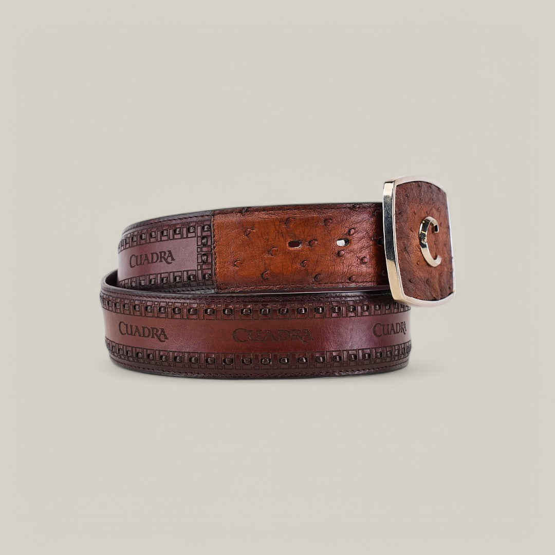 A coiled brown leather belt with intricate patterns showcases a silver rectangular buckle adorned with a decorative C emblem. CUADRA is embossed repeatedly, reminiscent of the craftsmanship in the Moroni Chestnut Ostrich Laser & Woven Belt - BC278. The plain background highlights its elegance.