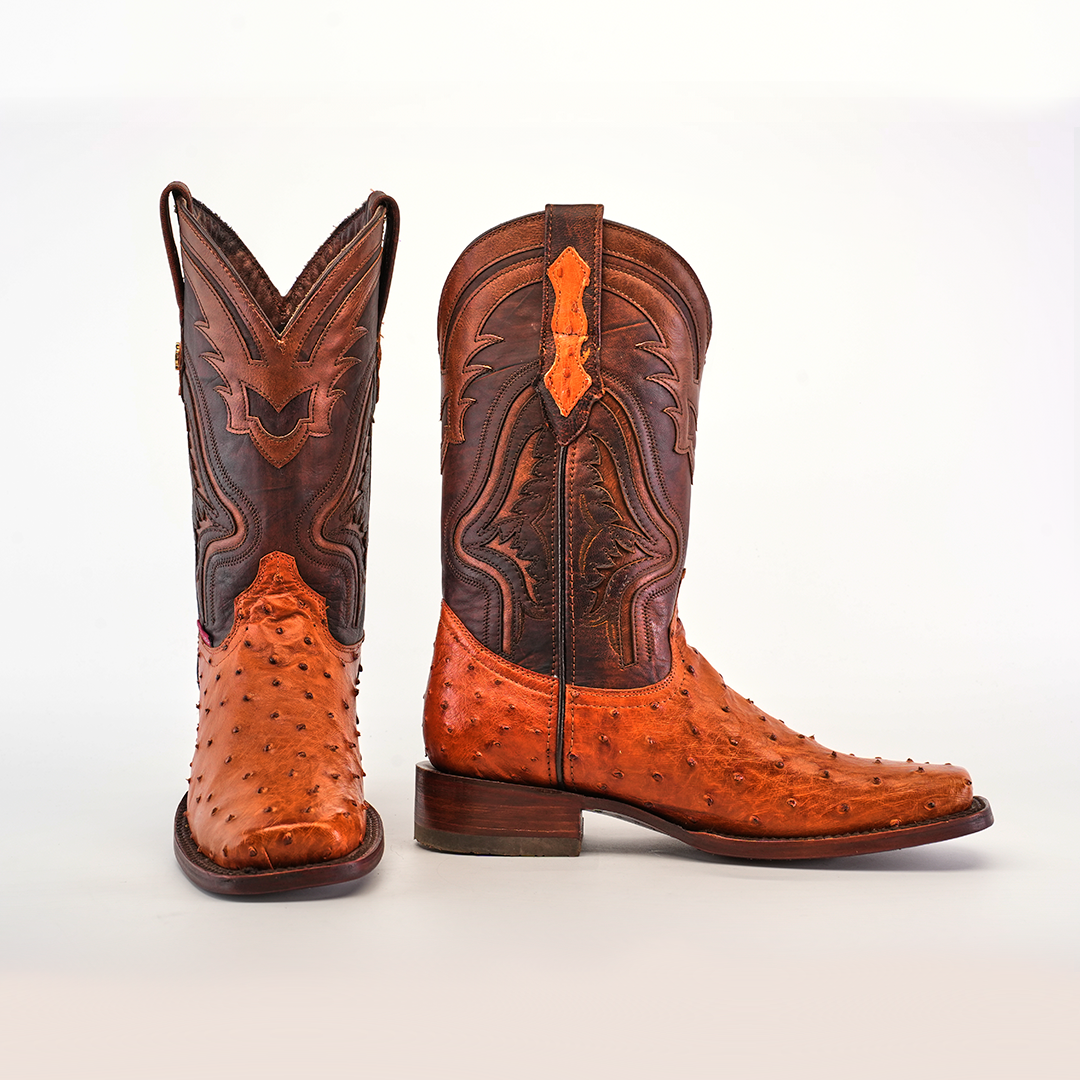 The Exotic Ostrich - Brandy - Rodeo Toe boots, crafted from textured brown ostrich leather with intricate embroidery, are displayed against a white background. One boot stands upright while the other tilts to showcase its sole, highlighting the exceptional craftsmanship.