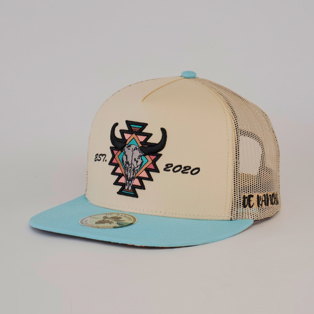 The De Rancho Cap Aztec Turquoise features a beige and blue trucker style with a mesh back, showcasing Western heritage. A geometric bull skull and EST. 2020 adorn the front, while DC Ranch in black on the side captures ranch culture.