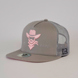 The De Rancho Cap Cassic Gray features a pink embroidered cowboy, symbolizing ranch culture, with 2020 stitched next to it. It includes a mesh back, a side logo patch labeled De Rancho, and a round sticker on the flat brim, merging style and tradition.