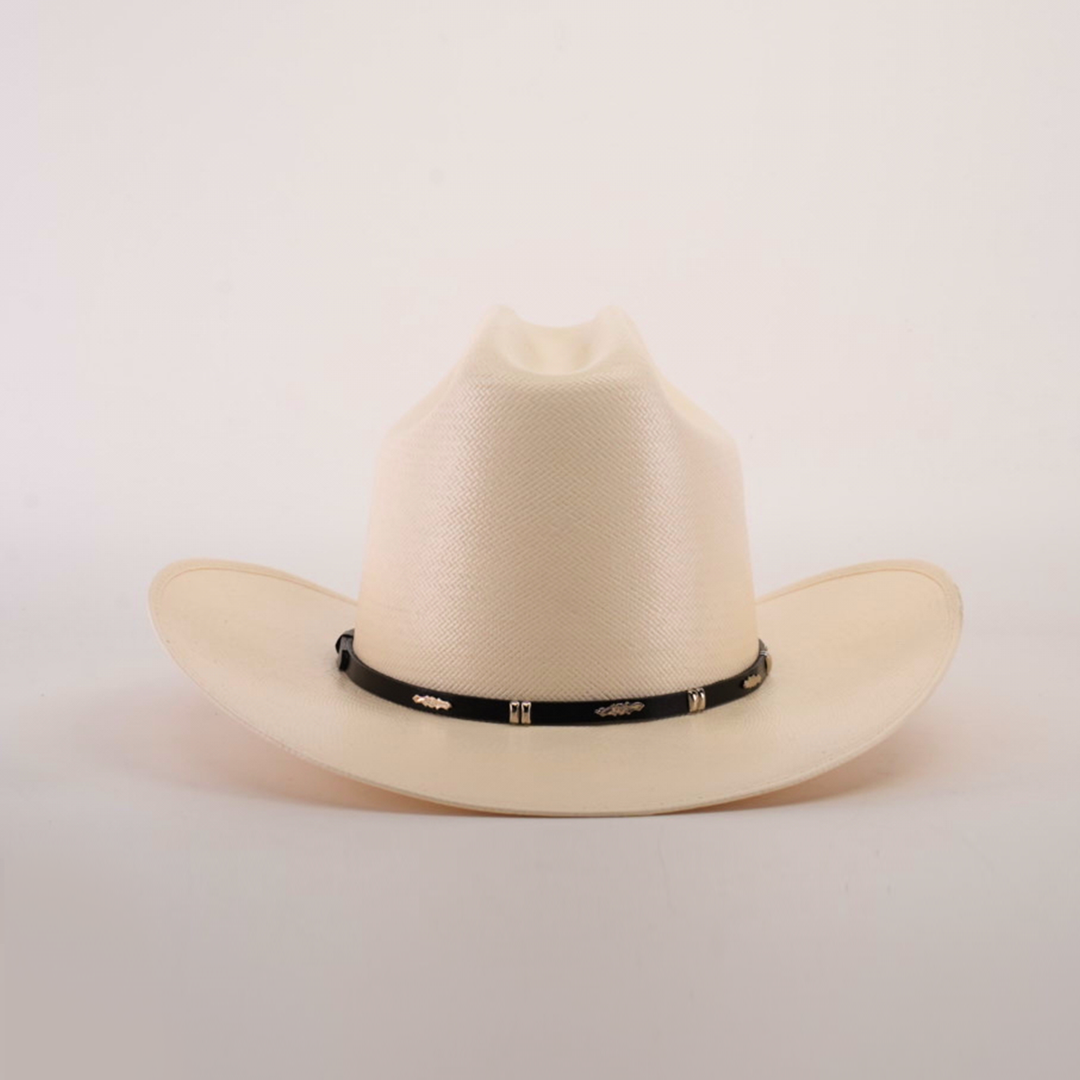 The 500X Dos Carnales Straw Hat is cream-colored with a wide brim and slightly pinched crown, highlighting its quality and durability. It has a black band adorned with decorative gold elements on a plain white background.