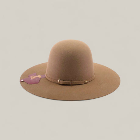 The 10x - Natural - TC Hats is a wide-brimmed, light brown felt western hat with a simple band around the crown, featuring a purple tag on the left brim. The hat is displayed against a plain beige background.