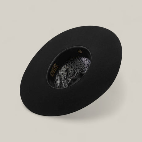 The 4X - Black - TC Hat is displayed upside down, showing its ornate patterned lining with gold lettering. Its shapeable crown is set on a light beige surface.