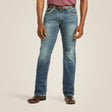 A person wearing M5 Slim Gambler Stackable Straight Leg Jean from Ariat, with a brown belt, stands with hands at their sides. They don brown leather shoes and a maroon shirt against a plain light beige background.