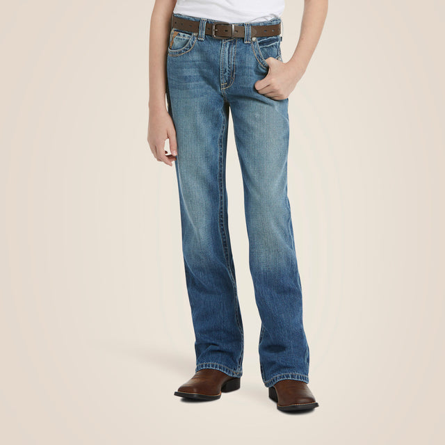 A person leans casually against a plain beige backdrop, wearing a white shirt, Ariat Boys B4 Boundary Dakota Medium Wash Relaxed Fit Boot Cut Jeans - 10018345, brown cowboy boots, and a matching brown belt with one hand in their pocket.