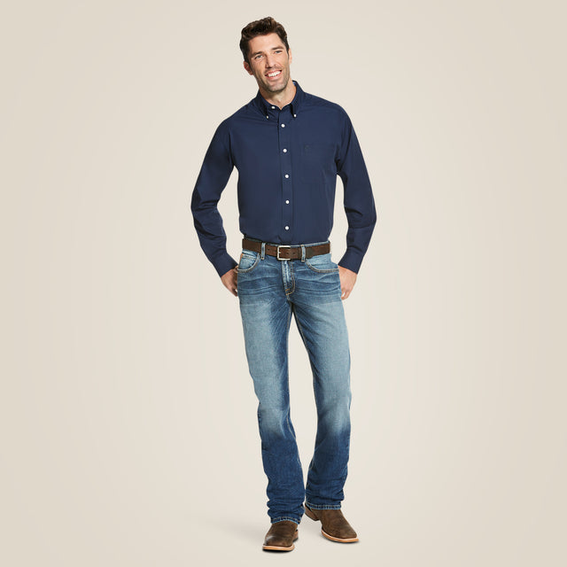 A man in a navy Wrinkle Free Solid Classic Fit Shirt stands smiling against a beige background, hands in pockets. Complemented by blue jeans, brown shoes, and a belt, his look showcases both style and wrinkle-resistant technology.