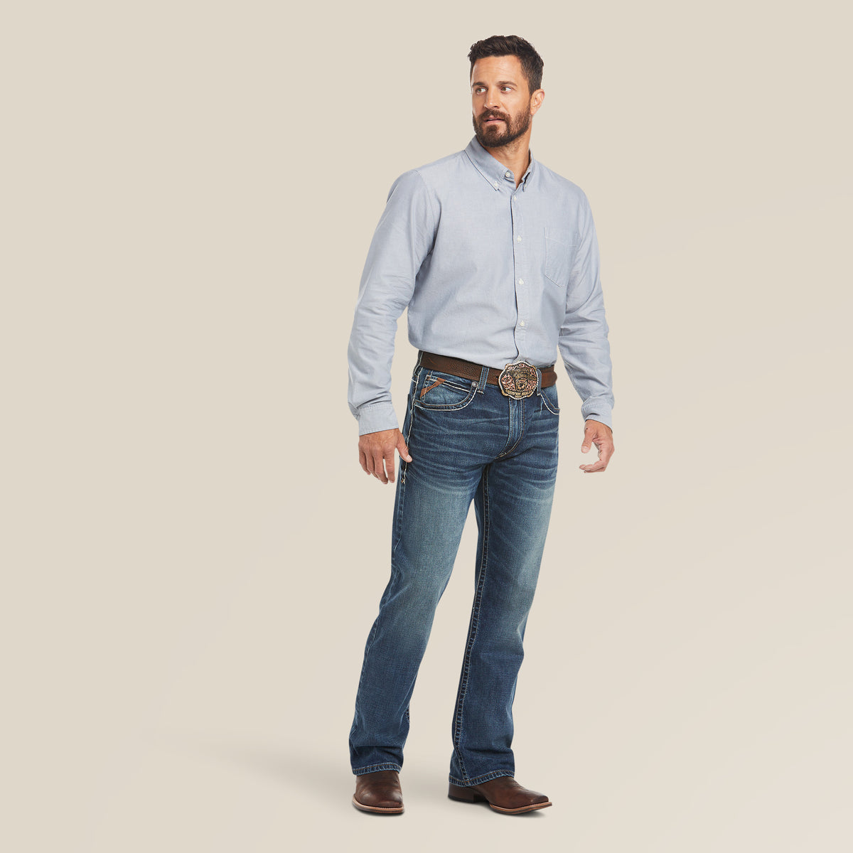 A bearded man stands against a plain background, wearing a light blue button-up shirt and M4 Low Rise Stretch Preston Boot Cut Jeans (Silverton - 10023455). His brown cowboy boots peek from the boot cut, complemented by a decorative belt with a large buckle. He gazes thoughtfully off to the side.