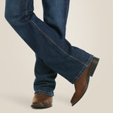 A person is casually standing with legs crossed, showcasing Ariat Boys B4 Legacy Dark Wash Relaxed Fit Boot Cut Jeans, paired perfectly with brown leather cowboy boots featuring a black heel against a neutral beige backdrop.