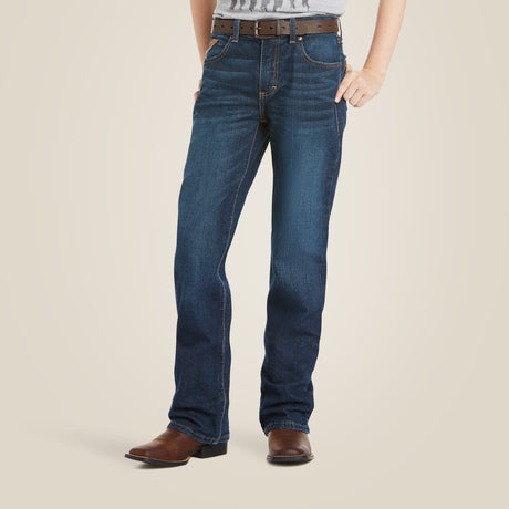 A person in Ariat Boys B4 Legacy Dark Wash Relaxed Fit Boot Cut Jeans (10027675), a brown belt, brown cowboy boots, and a tucked-in gray shirt stands against a plain background with hands in pockets.