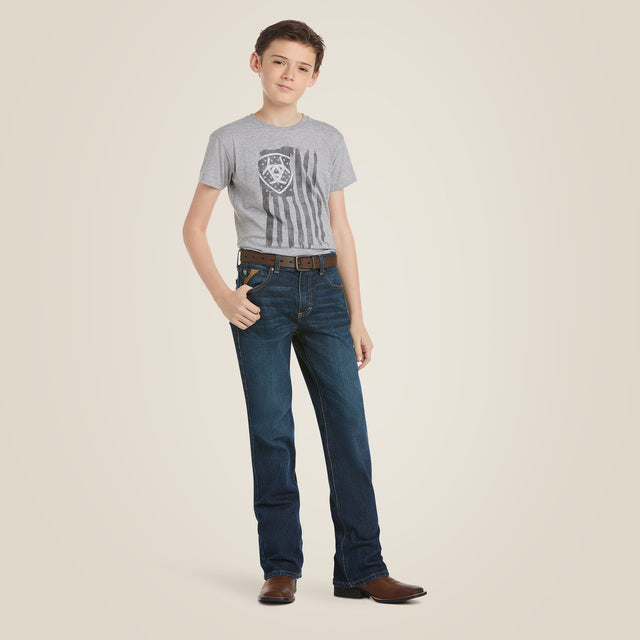 A young person wearing a gray T-shirt with a shield and stripes design, Ariat Boys B4 Legacy Dark Wash Relaxed Fit Boot Cut Jeans, and brown cowboy boots stands confidently with one hand in their pocket against a neutral background.