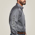 A man wears the Team Logo Twill Classic Fit Shirt Gray - 10037479, showcasing ARIAT embroidered on the sleeve. Complementing his jeans and brown belt, the structured collar highlights his beard against a simple, light background.