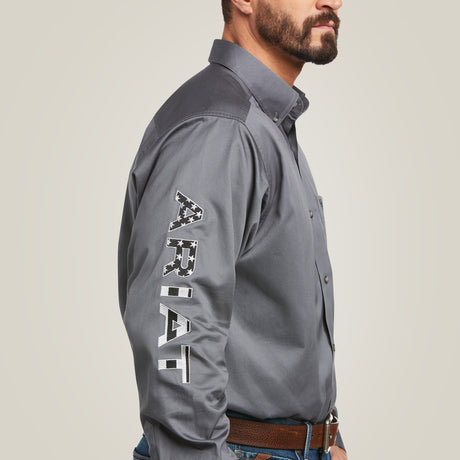 A man wears the Team Logo Twill Classic Fit Shirt Gray - 10037479, showcasing ARIAT embroidered on the sleeve. Complementing his jeans and brown belt, the structured collar highlights his beard against a simple, light background.