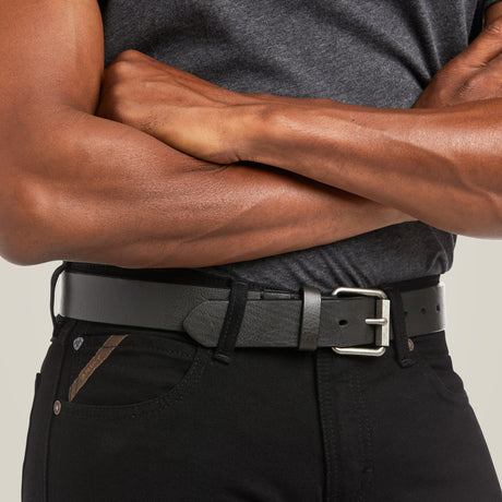 A modern cowboy stands confidently in a gray shirt and M7 Slim Legacy Straight Jean - Black (10037890), arms crossed. His black leather belt with a metallic buckle complements the premium denim, accentuated by the pocket’s distinctive small brown stripe detail.