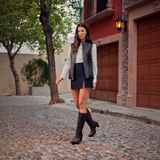 A woman walks confidently down a cobblestone street, her Maple Zipper & Stud & Laser - Round Toe boots complementing a black vest over a light shirt and black skirt. A red building and trees serve as the backdrop, with a warm lantern glow epitomizing luxury craftsmanship in every detail.