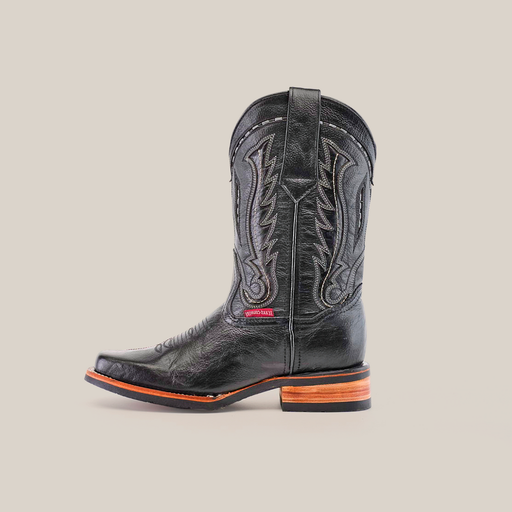 The Barcelona Black boot, made from premium cowhide, showcases decorative stitching and a rodeo toe. It pairs a stacked brown and black heel with a slip-resistant sole. Set against a plain white background, the boot features a subtle side label.