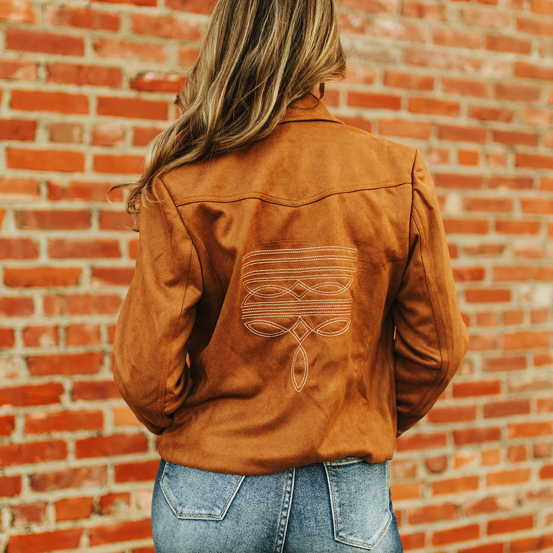 A person with long hair stands against a red brick wall wearing the LB Camel Suede Western Stitch Blazer, beautifully adorned with embroidered geometric patterns. The luxury jacket complements the rustic backdrop and adds an upscale touch to their attire.