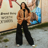 A modern woman smiles broadly in front of a mural, donning the LB Camel Western Stitch Bomber JO241-CML and black wide-leg pants. The bombers western details complement the cowboy mural as her hands rest confidently on her jacket.