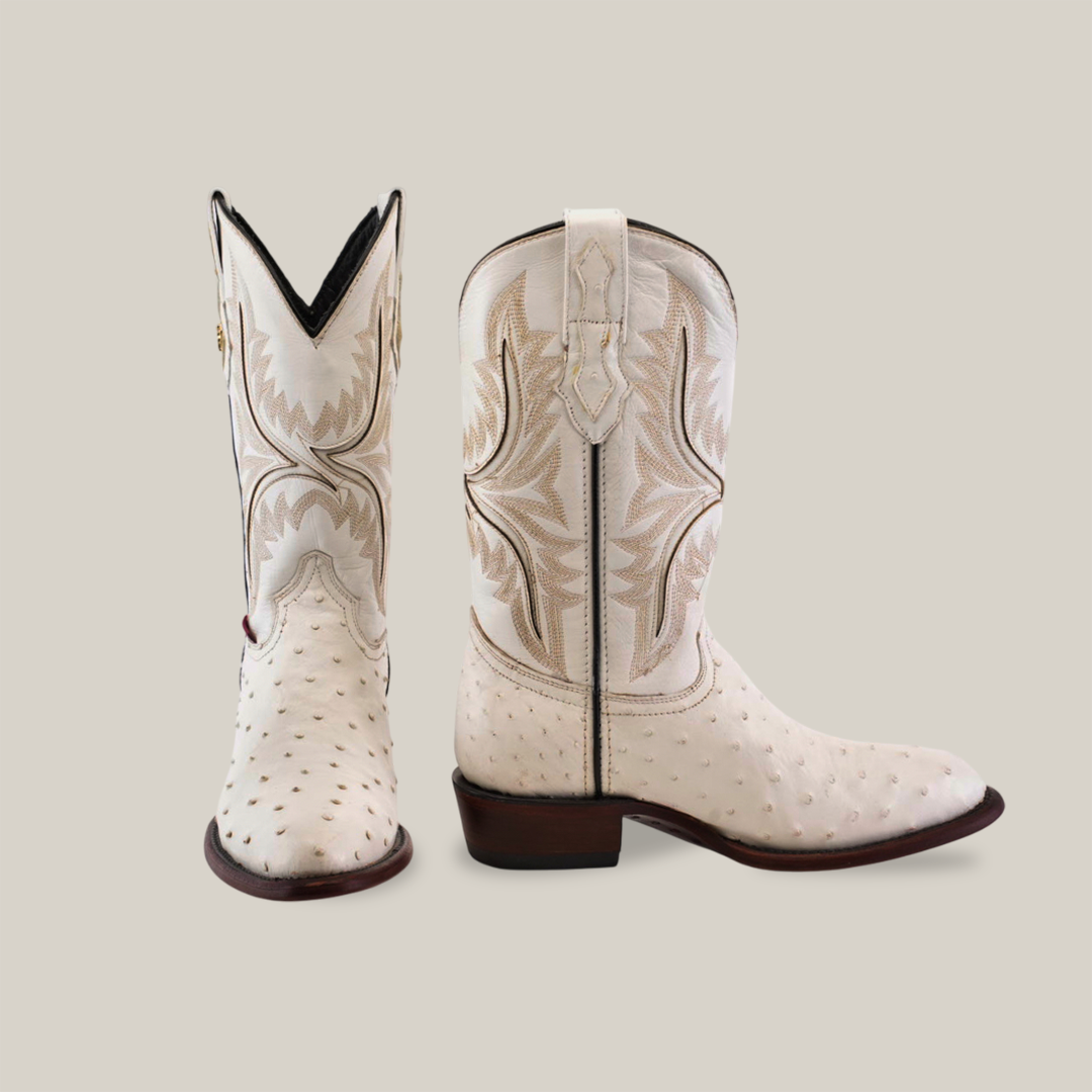 The Exotic Full Quilt Ostrich boots in white feature intricate brown embroidery. One boot faces forward, showcasing a detailed V-shaped top, while the side-facing boot highlights the decorative pattern. These handmade boots flaunt wooden heels and an authentic ostrich leather texture.