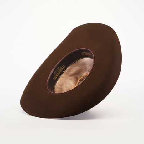 The 6X Brazil Brown felt cowboy hat is angled to reveal its inside brim and decorative lining with a patterned label. It casts a subtle shadow, highlighting its depth and texture against a light background, showcasing its Western heritage.