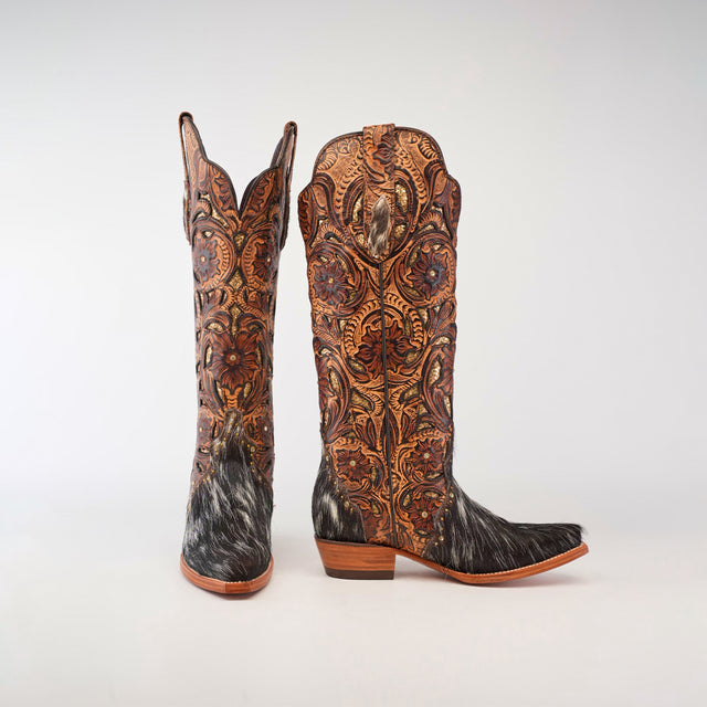 The Handtooled Cowhide Pinto boots are intricately designed womens cowboy boots featuring ornate tooled leather patterns and fur accents. These handcrafted boots boast a tall shaft, stacked heel, and blend brown hues with detailed craftsmanship against a light gray background.