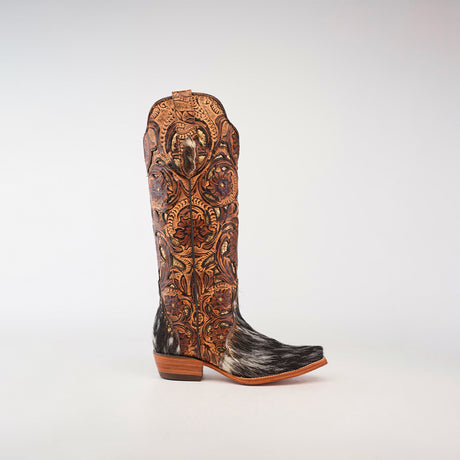 The Handtooled Cowhide Pinto - Tall Shaft - Snip Toe boots feature intricate brown leather tooling and dark speckled hair-on-hide details, set against a light gray background. Handcrafted with precision, these boots exude rustic elegance.