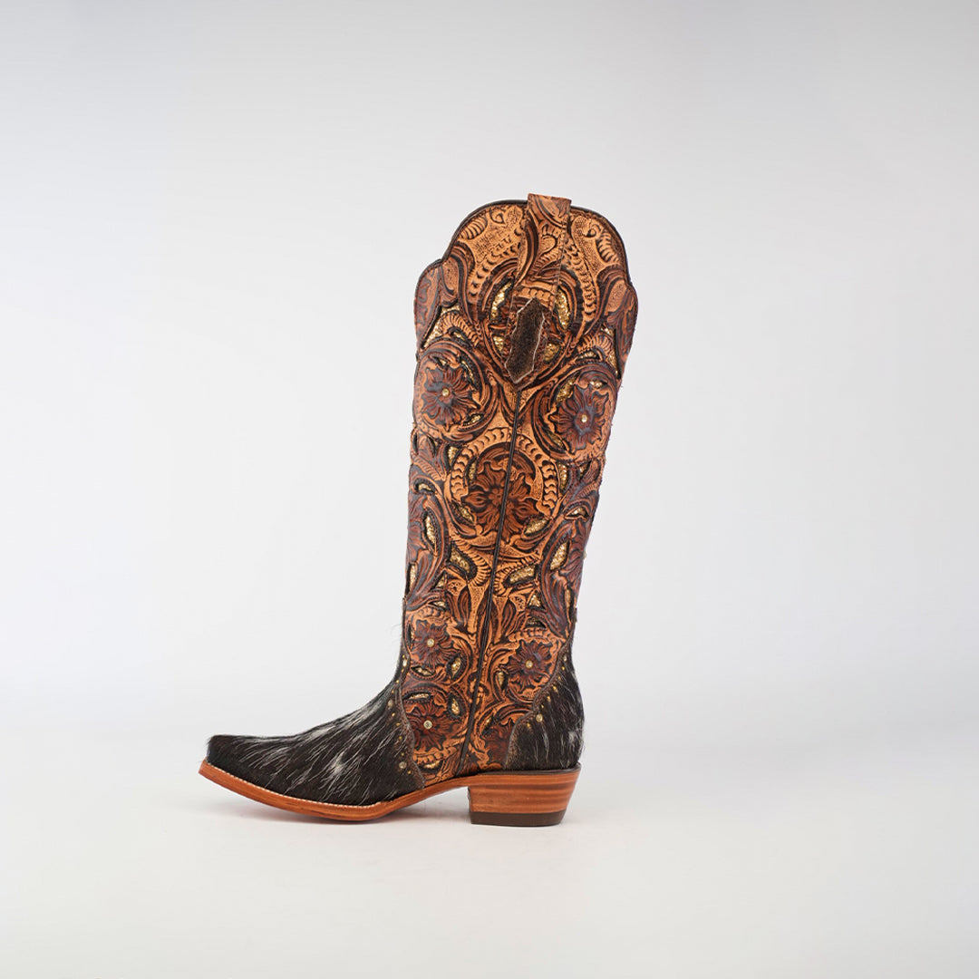 The Handtooled Cowhide Pinto boot is a tall, handcrafted womens boot with intricate brown and orange stitching on the shaft and a black textured cowhide lower, all set against a white background.