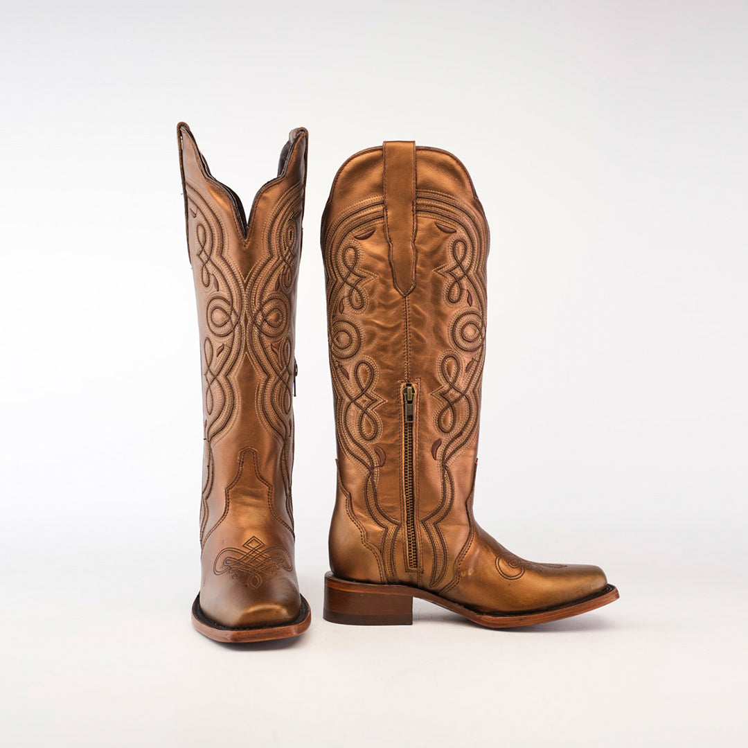 The Eden Copper Tall Shaft boots, with a narrow square toe, showcase intricate brown leather embroidery and a side zipper. One boot stands upright while the other is angled side-on, highlighting their luxury craftsmanship and block heel design.