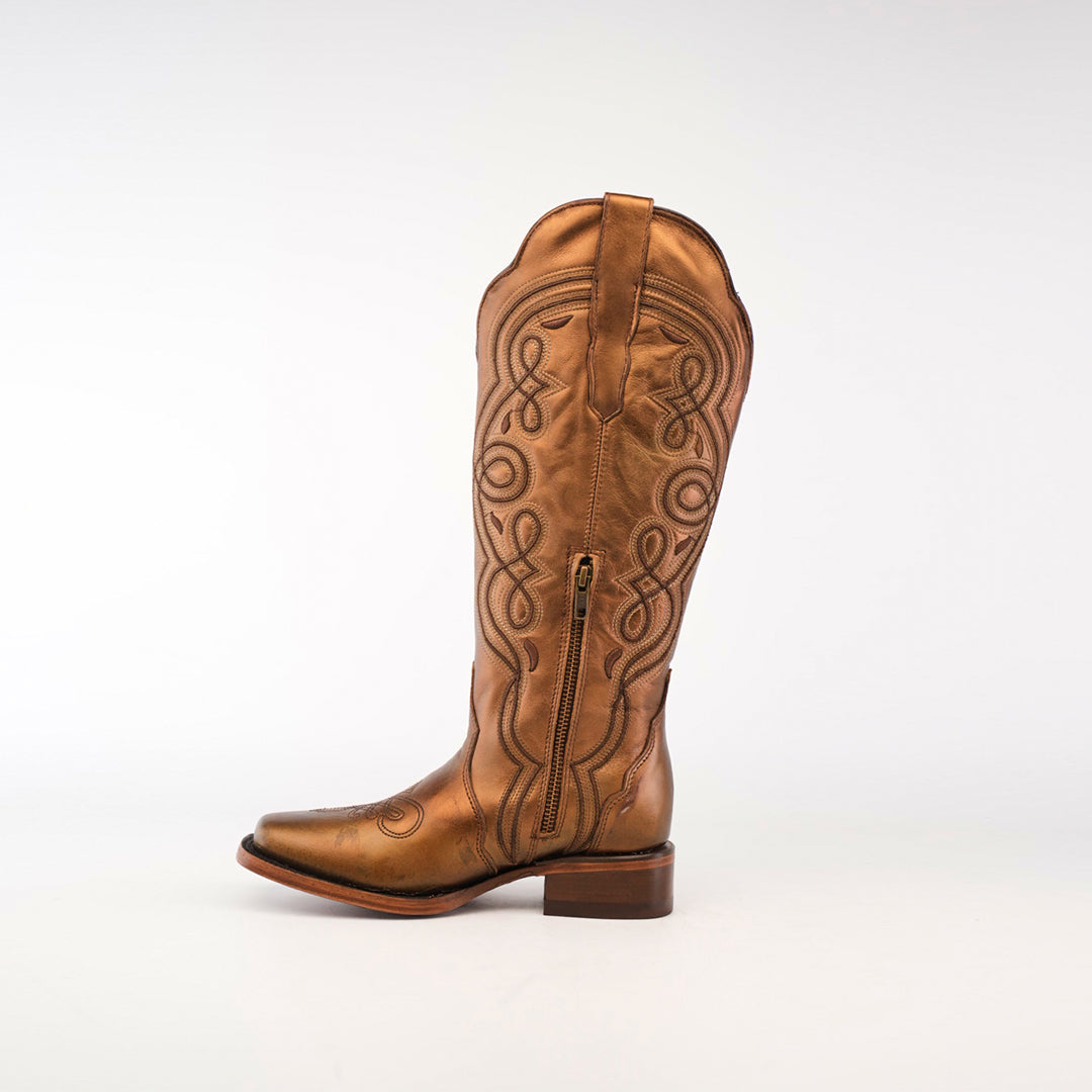 The Eden Copper Tall Shaft boot in premium brown leather features intricate swirl embroidery, a low heel, and a side zipper. It stands upright against a plain white background, highlighting luxury craftsmanship.