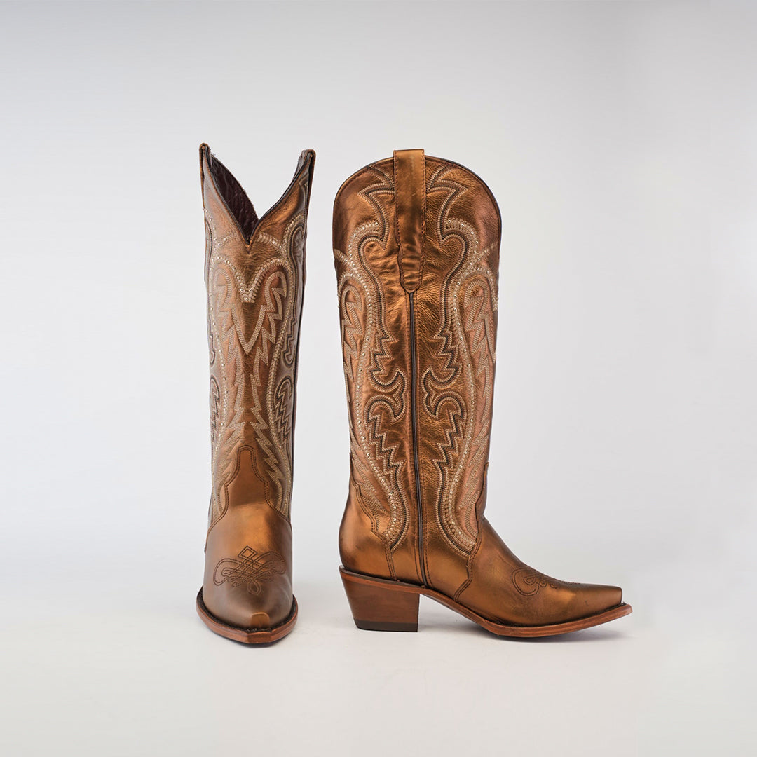 The Nihan Copper - Tall Shaft - Snip Toe boots, featuring intricate shaft stitching and pointed toes, highlight traditional craftsmanship. One boot stands upright while the other tilts slightly, showcasing their handcrafted excellence against a plain light gray background.