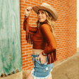 A woman outdoors in a boho chic style wears the LB Camel Crop Long Sleeve Fringe TO486-SCML and a denim skirt. She leans against a brick wall with a straw cowboy hat, her tattoo visible as she gazes into the distance with a slight smile.