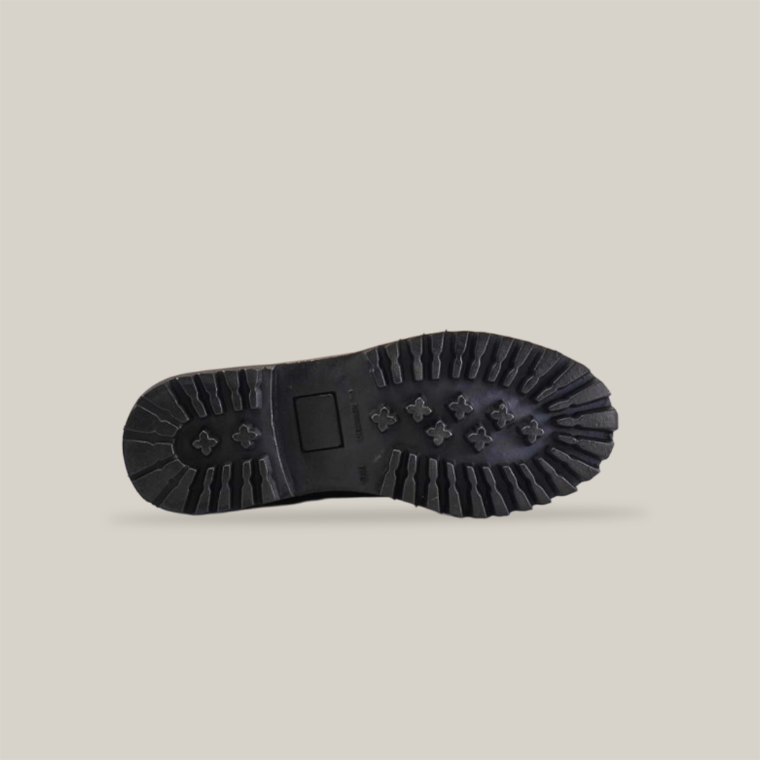 The image shows a black shoe sole with deep, rugged treads and various rectangular and circular indents on a plain light background, highlighting the tough tractor sole design and grip pattern inspired by the Brown Roper - Tractor Sole - Soft Toe.