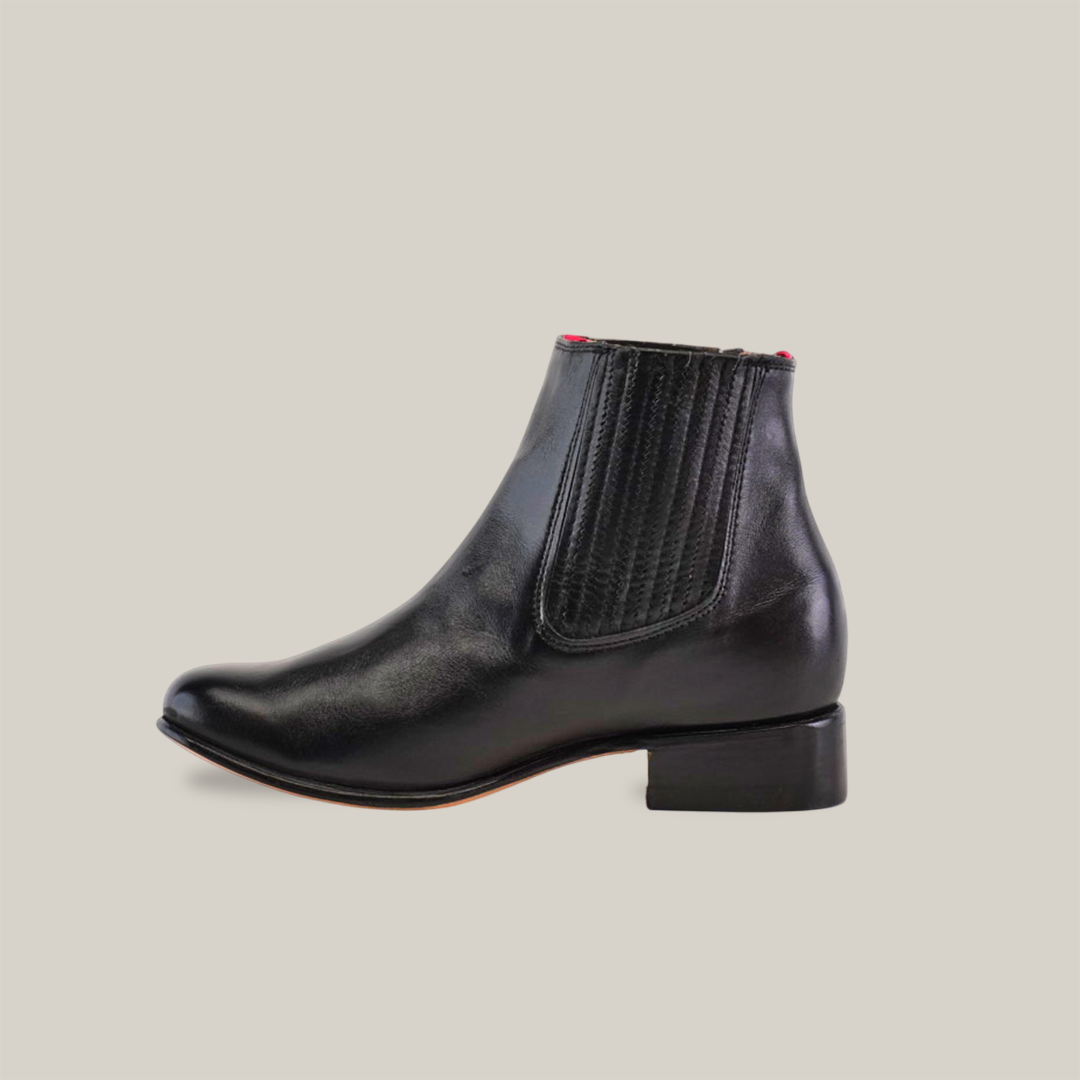 The Napa Black - Round Toe is a black leather ankle boot with an elastic side panel, a short block heel inspired by cowboy fashion, and features a red tag and branding. Its visible light-colored sole adds modern flair to the classic design.