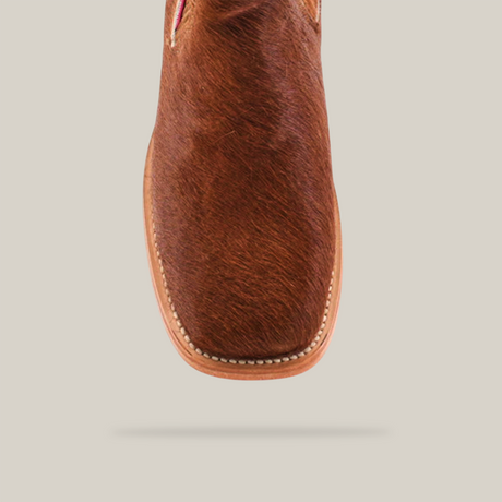 A brown, textured Cowhide Hair Camel Square Toe shoe is set against a white background. It exudes Western flair with its cowhide hair-like finish and detailed stitching along the soles edge.