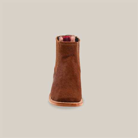 Front view of the Cowhide Hair Camel Square Toe boot, featuring a brown suede finish, rounded toe, and flat sole. It includes a touch of fur on top and a visible small red tag inside. The scene is set against a plain light background, echoing traditional Western footwear styles.