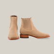 The Prime Suede Pearl Square Toe boots are handcrafted in beige suede with a wooden heel. The left boot faces forward, the right showcases the elastic side panel, all against a plain white backdrop.