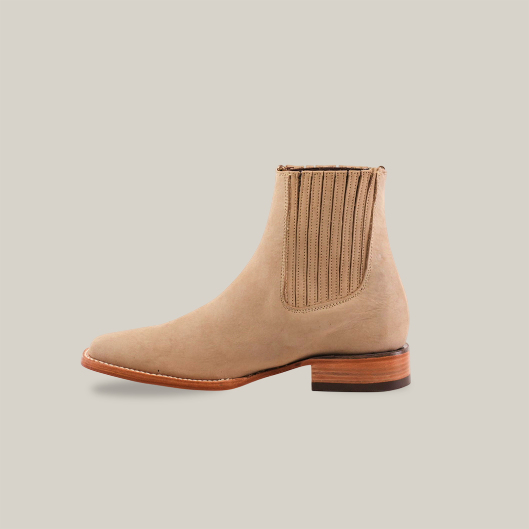 The Prime Suede Pearl Square Toe boots, handcrafted in elegant beige suede with a wooden heel and side elastic panels, capture timeless style. Their cowboy-inspired design stands out against a plain white background.