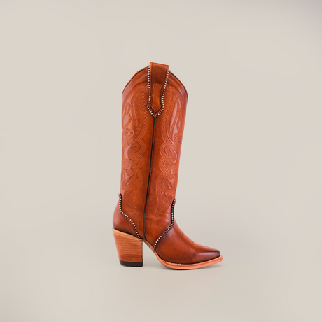 The Linda Tall Shaft Tan - J Toe boot from the Platinum Collection is displayed against a simple light backdrop. Made from premium leather, it features intricate stitching, a tall shaft, pointed toe, and chunky heel.