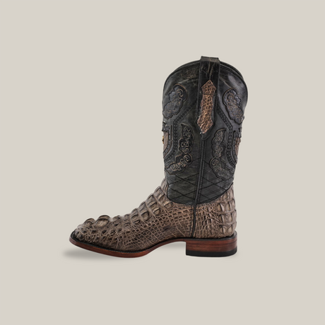 The Exotic American Alligator Head - Gray - Square Toe cowboy boot, featuring intricate designs on the shaft and an alligator texture on the foot section, is handcrafted from luxurious gray alligator leather and boasts a sturdy wooden heel, displayed against a plain, light background.