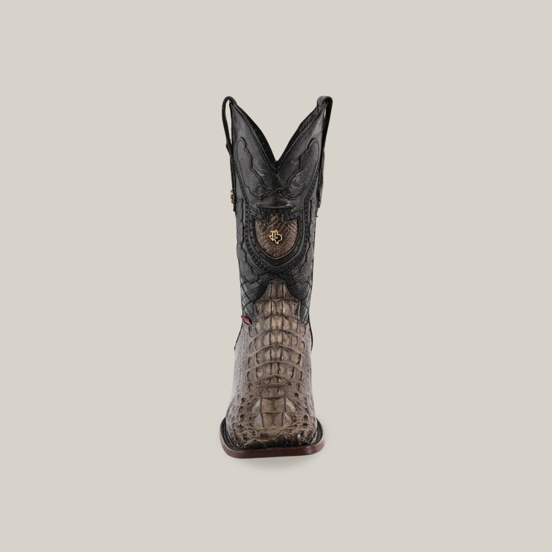 A front view of the Exotic American Alligator Head boot in gray with a square toe showcases a textured, crocodile-patterned lower section and intricate black leather designs on the upper, featuring a decorative emblem against a neutral beige background.