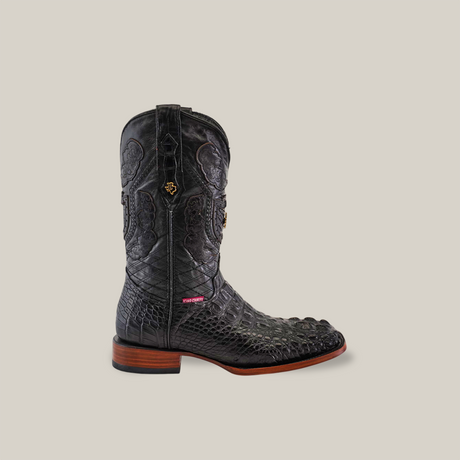 The Exotic American Alligator Head boot in black showcases a textured, crocodile-like pattern with gold detailing. It features a striking square toe, brown wooden heel and toe, and a red tag near the sole, set against a beige background for an authentic cowboy boot look.