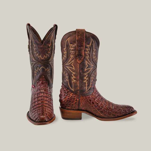 The Exotic Caiman Hornback boots in brown showcase intricate stitching and textured leather, crafted from authentic caiman leather. One boot stands upright while the other tilts slightly, highlighting the detailed patterns on both the shaft and foot.