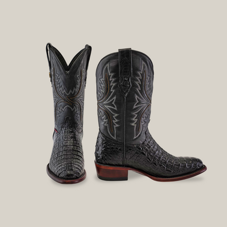 The handcrafted black cowboy boots, made from exotic Caiman Hornback leather, feature intricate stitching and textured surfaces. One boot faces forward with a round toe, while the other shows the heel and decorative patterns.