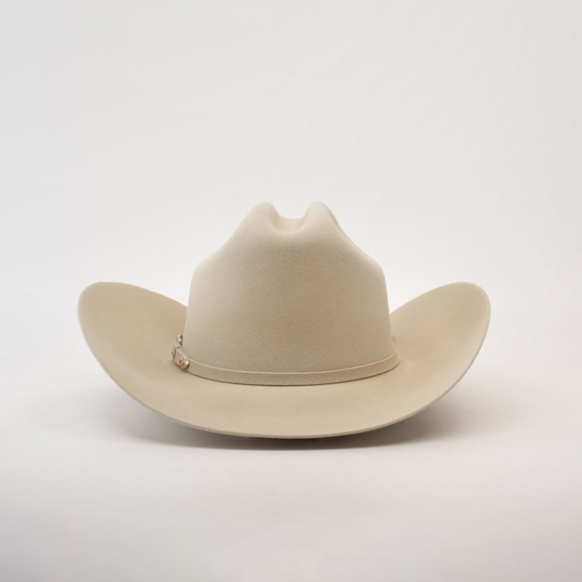 The 6X Real Belly is a beige fur felt cowboy hat with a wide brim and crease, set against a white background. It features a thin band with small decorative elements on the side, crafted from premium furs.