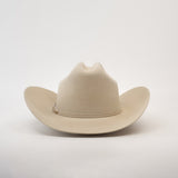 The 6X Real Belly is a beige fur felt cowboy hat with a wide brim and crease, set against a white background. It features a thin band with small decorative elements on the side, crafted from premium furs.