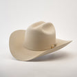 The 500X Superior Belly, a beige cowboy hat made from 100% belly beaver fur, features a wide brim and a decorative band with a metallic embellishment. Displayed on a smooth light gray surface, it is slightly tilted to highlight its premium craftsmanship.