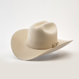 The 1000X Imperial Belly hat is a beige cowboy style with a wide brim, adorned with a decorative band featuring a small metallic emblem, set against a plain white background.